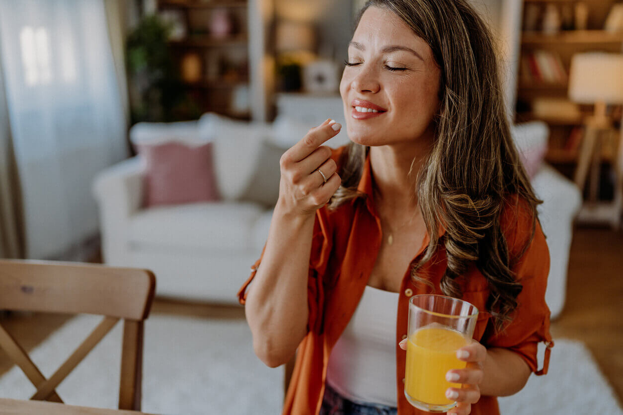 Mulher sorridente prestes a tomar um comprimido enquanto segura um copo de suco de laranja, representando os cuidados com a saúde durante o inverno e a importância de equilibrar probióticos e antibióticos.